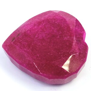 Könnte beinhalten: Ein facettierter, herzförmiger Edelstein in leuchtendem Magenta. Der Stein hat mehrere Facetten, die das Licht einfangen und einen funkelnden Effekt erzeugen. Der Stein ist vor einem weißen Hintergrund platziert, der seine Farbe und Form hervorhebt.