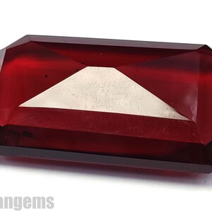 May include: A faceted, rectangular gemstone in a deep red colour. A white, triangular shape is visible in the centre of the gem. The gem is set against a white background. The word "Rohangems" is visible at the bottom left.