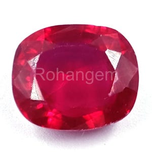 Puede incluir: Una gema de rubí facetada, tallada en forma de cojín, de un tono rojo intenso. La gema tiene el texto "Rohangem" impreso en su superficie. El rubí se encuentra sobre un fondo blanco, resaltando su color y claridad.