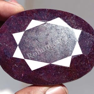 May include: A large, oval-shaped, faceted ruby gemstone with a star-shaped pattern in white. The ruby has a deep red colour with a slightly purple hue.