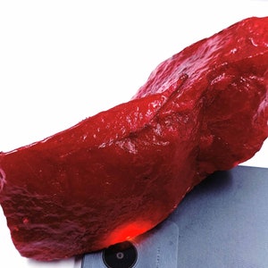 May include: A large, rough, red gemstone. The gemstone is irregularly shaped and has a glossy finish.