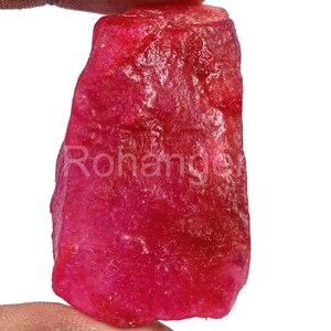 May include: A raw, uncut ruby gemstone in a deep red colour. The surface has a textured appearance, and the word "Rohange" is visible on the stone. The ruby is held between two fingers against a white background.