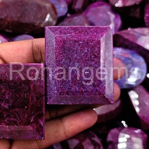 May include: A close-up of a hand holding two large, square-shaped, purple gemstones. The gemstones are faceted and have a deep, rich color. The gemstones are surrounded by other purple gemstones of various shapes and sizes.