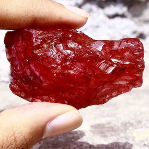 Peut inclure: Une grosse pierre précieuse rouge brute avec une structure cristalline. La pierre précieuse est tenue dans une main.