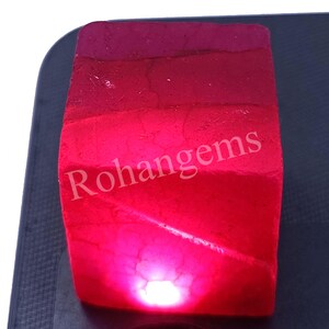 May include: A deep red gemstone with a rough, faceted surface. The gemstone is illuminated from below, casting a bright red glow. The text "Rohangems" is visible in the image.