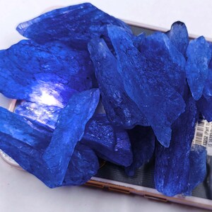 May include: A collection of raw, deep blue kyanite crystals. The crystals have a rough, textured surface and varying shapes, with sharp edges and a natural, unpolished appearance. The kyanite is arranged on a light-colored surface.