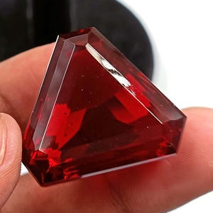 May include: A deep red gemstone with a triangular shape and many facets. The gemstone is cut in a unique style, with a sharp point at the top and a flat base.