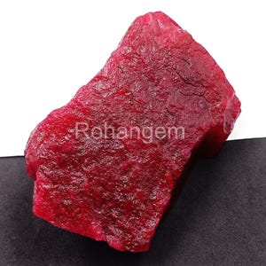 Può includere: Una gemma di rubino grezza, non tagliata, di colore rosso intenso. La superficie ha un aspetto strutturato. La parola "Rohangem" è stampata sulla superficie della gemma. La gemma è esposta su uno sfondo bianco e nero.