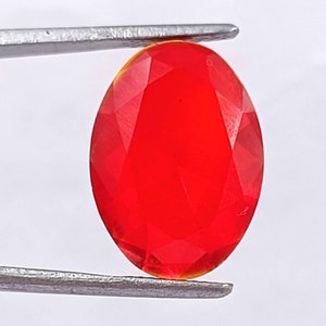 May include: A bright red oval-shaped gemstone with a faceted cut. The gemstone is held by a pair of tweezers.