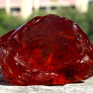 Puede incluir: Una gema grande, cruda, translúcida y de color rojo intenso con una forma irregular y dentada, y una superficie brillante y reflectante. La piedra muestra varios tonos de carmesí y escarlata con bordes afilados y fracturas naturales.