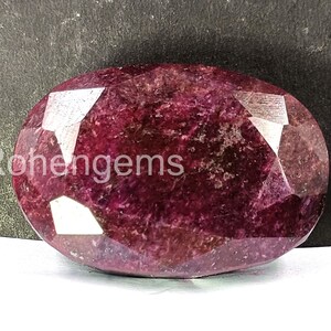 May include: A faceted, oval-shaped ruby gemstone. The gem is a deep, rich red color with a polished surface. The text "Rohengems" is visible on the left side of the gemstone.