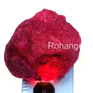 Puede incluir: Una gema de rubí en bruto, sin cortar, de color rojo intenso. La superficie tiene un aspecto texturizado y la palabra "Rohang" está impresa en la superficie. La gema se coloca sobre un dispositivo de medición plateado.