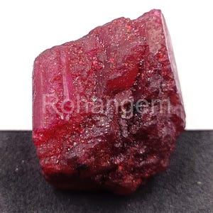 May include: A raw, deep red ruby gemstone with a textured surface. The word "Rohangem" is visible on the surface. The ruby displays a rich, saturated colour and a natural, unpolished appearance, showcasing its raw beauty.
