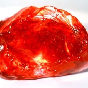 Puede incluir: Una gema rojo-naranja translúcida y vibrante con una superficie rugosa e irregular. La gema tiene un aspecto brillante, reflejando la luz y mostrando su estructura interna. La forma general es una piedra grande sin cortar, probablemente un espécimen mineral en bruto.