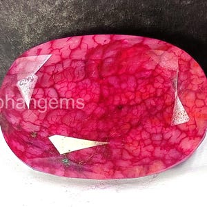 May include: Oval-shaped, faceted gemstone with a deep red hue and a unique, textured pattern. The gemstone has a few white, triangular facets. The text "Rohangems" is visible on the left side of the gemstone.