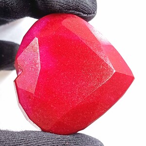 May include: A deep red heart-shaped gemstone with a faceted cut. The gemstone has a smooth, polished surface.