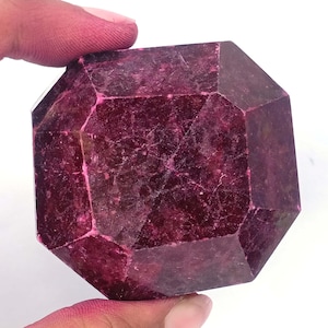 Puede incluir: Una gema facetada, de color burdeos intenso, con forma geométrica. La gema tiene una superficie pulida con patrones internos visibles y un color rico y saturado. La piedra está sostenida por una mano, mostrando su tamaño y forma.