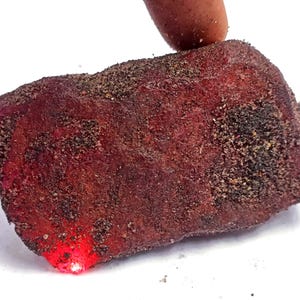 Puede incluir: Una gema en bruto, de color rojo intenso, con una superficie texturizada y una capa de partículas oscuras. El borde inferior de la piedra brilla con una luz roja brillante, lo que sugiere que es translúcida. La piedra está sujeta por un dedo.