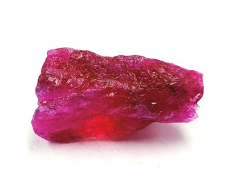 May include: A raw, unpolished ruby gemstone with a deep, vibrant magenta and red color. The stone features a jagged, irregular crystalline structure with translucent sections that catch the light, showcasing natural facets and rough, textured surfaces.
