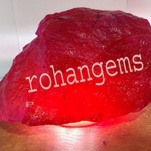 May include: A large, rough, red gemstone with the text "rohangems" on it. The gemstone is sitting on a wooden base.