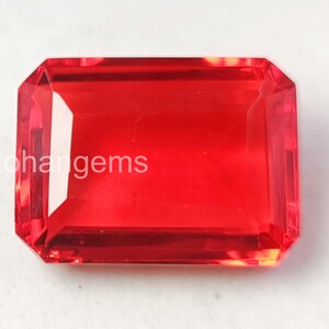 May include: A faceted, rectangular red gemstone with the text "Rohangems" visible. The gem has a vibrant, transparent appearance and a polished finish. The shape is an emerald cut, with beveled edges.
