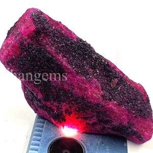 May include: A large, rough-cut ruby gemstone with a vibrant magenta hue, interspersed with dark, almost black, mineral inclusions. The stone is irregularly shaped and appears to be unpolished, showcasing its natural texture. The word "angems" is visible on the stone.