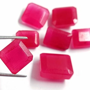 May include: Seven pink gemstone cabochons, cut into square shapes, with a slightly rounded edge. The gemstones are displayed on a white surface, with one being held by tweezers.