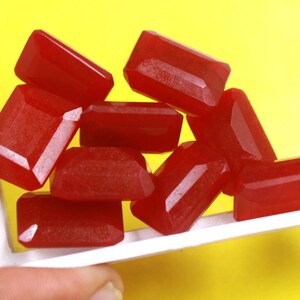 Pu&ograve; includere: Un gruppo di otto cabochon di pietre preziose rosse di forma rettangolare. Le pietre preziose sono esposte in una scatola bianca su uno sfondo giallo.