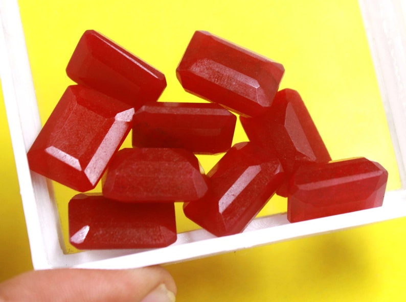 Pu&ograve; includere: Un gruppo di otto cabochon di pietre preziose rosse di forma rettangolare. Le pietre preziose sono esposte in una scatola bianca su uno sfondo giallo.