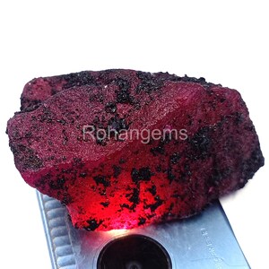 May include: A rough, uncut ruby gemstone with a deep red hue and dark, speckled inclusions. The surface of the stone is textured, and the word "Rohangems" is visible on the surface. The gemstone is displayed on a light-colored surface.