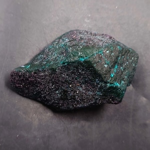 May include: A dark green and purple rock with a rough, textured surface. The rock has a speckled appearance with small, light blue flecks.