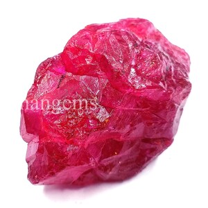 May include: A rough, uncut ruby gemstone with a deep red hue. The surface is textured and irregular, with visible crystalline structures. The word "nangems" is visible on the surface. The gemstone is set against a white background.