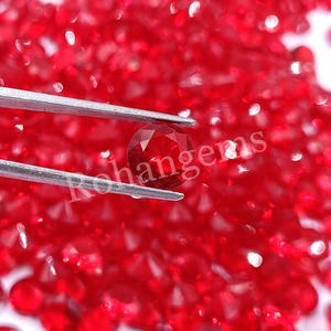 Puede incluir: Primer plano de una sola piedra preciosa roja sostenida por unas pinzas sobre un fondo de piedras preciosas rojas sueltas. La piedra preciosa es redonda y facetada, con un alto grado de brillo.