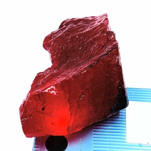 May include: A large, rough-cut, translucent red gemstone with a textured surface. The gemstone has a deep, rich colour and appears to be freshly cut. The stone is set against a white background.