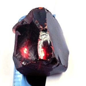 May include: A dark, faceted gemstone in a deep burgundy hue. The stone has a rough, natural texture with areas of high reflectivity, and a bright red glow emanating from within. The gemstone is held by a blue support.