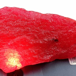 May include: A large, rough, red gemstone with a shiny, translucent surface. The gemstone is sitting on a black surface.