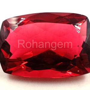 May include: A faceted, rectangular-cut gemstone in a vibrant red hue. The gem has a polished surface with multiple facets that reflect light. The text "Rohangem" is visible on the surface of the gem.