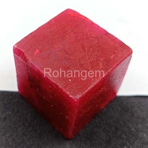 May include: A vibrant red, cube-shaped gemstone with the text "Rohangem" on its surface. The gemstone has a slightly rough texture and appears to be a raw, uncut stone. The color is a deep, saturated red.