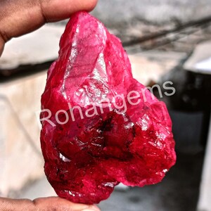 May include: A large, rough, red ruby gemstone. The ruby is a deep, rich red colour and has a natural, unpolished finish. The ruby is being held in a hand, and the word "Rohangems" is visible in the background.