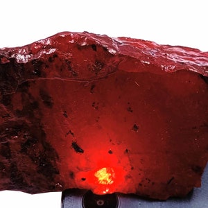 May include: A large, rough, red gemstone with a deep red hue. The gemstone is illuminated from below, highlighting its internal structure.