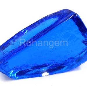 May include: A vibrant, translucent blue gemstone with a rough, irregular shape. The gem has a glossy surface and a deep, saturated color, reflecting light. The text "Rohangem" is visible on the surface.