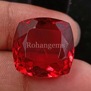 Puede incluir: Una piedra preciosa de color rojo intenso con talla cojín y muchas facetas. La piedra preciosa está tallada en forma cuadrada con esquinas redondeadas. La piedra preciosa refleja la luz y tiene un acabado brillante.