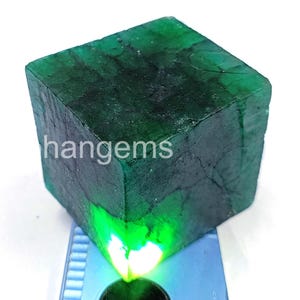 May include: A large, raw emerald gemstone, cut into a cube shape. The gem is a deep, rich green colour with visible internal textures and the text "hangems" on the side. The stone is displayed on a blue surface with a bright light shining through.