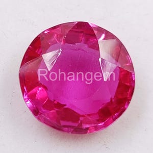 Puede incluir: Una gema magenta facetada y de corte redondo con el texto "Rohangem" en el centro. La gema tiene múltiples facetas que reflejan la luz, creando un efecto brillante. La piedra preciosa es de un tono rosa vibrante.