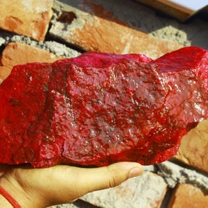 May include: A large, rough, red ruby gemstone. The ruby is a deep, rich red color and has a slightly translucent appearance.