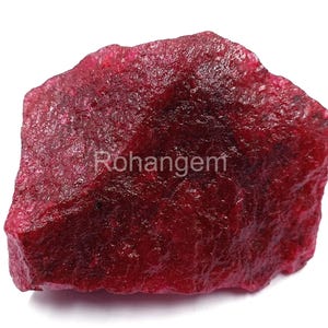 Puede incluir: Una gema de rubí en bruto, sin cortar, de color rojo intenso. La superficie tiene una apariencia texturizada, y la palabra "Rohangem" es visible. La gema se muestra sobre un fondo blanco.