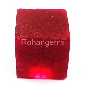 May include: A deep red, square-shaped gemstone with a rough, textured surface. The gemstone is illuminated from below, creating a bright red glow. The text "Rohangems" is visible on the surface of the gemstone.