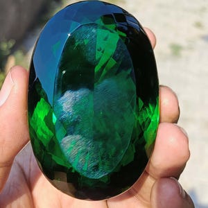 Può includere: Una grande gemma ovale sfaccettata, di un profondo colore verde smeraldo. La gemma è tenuta in mano, mostrando le sue superfici lucide e i riflessi interni. Le sfaccettature creano un gioco di luce, evidenziando la chiarezza e la brillantezza della gemma.