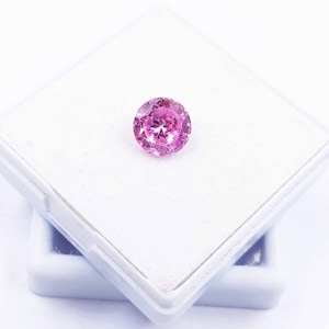 Può includere: Una pietra preziosa rotonda rosa con molte sfaccettature, posta in una scatola di presentazione quadrata bianca.