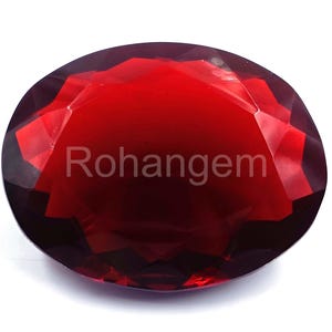 May include: A faceted, oval-shaped, deep red gemstone. The gem has a polished surface with multiple facets that reflect light. The text "Rohangem" is visible on the surface of the gem.